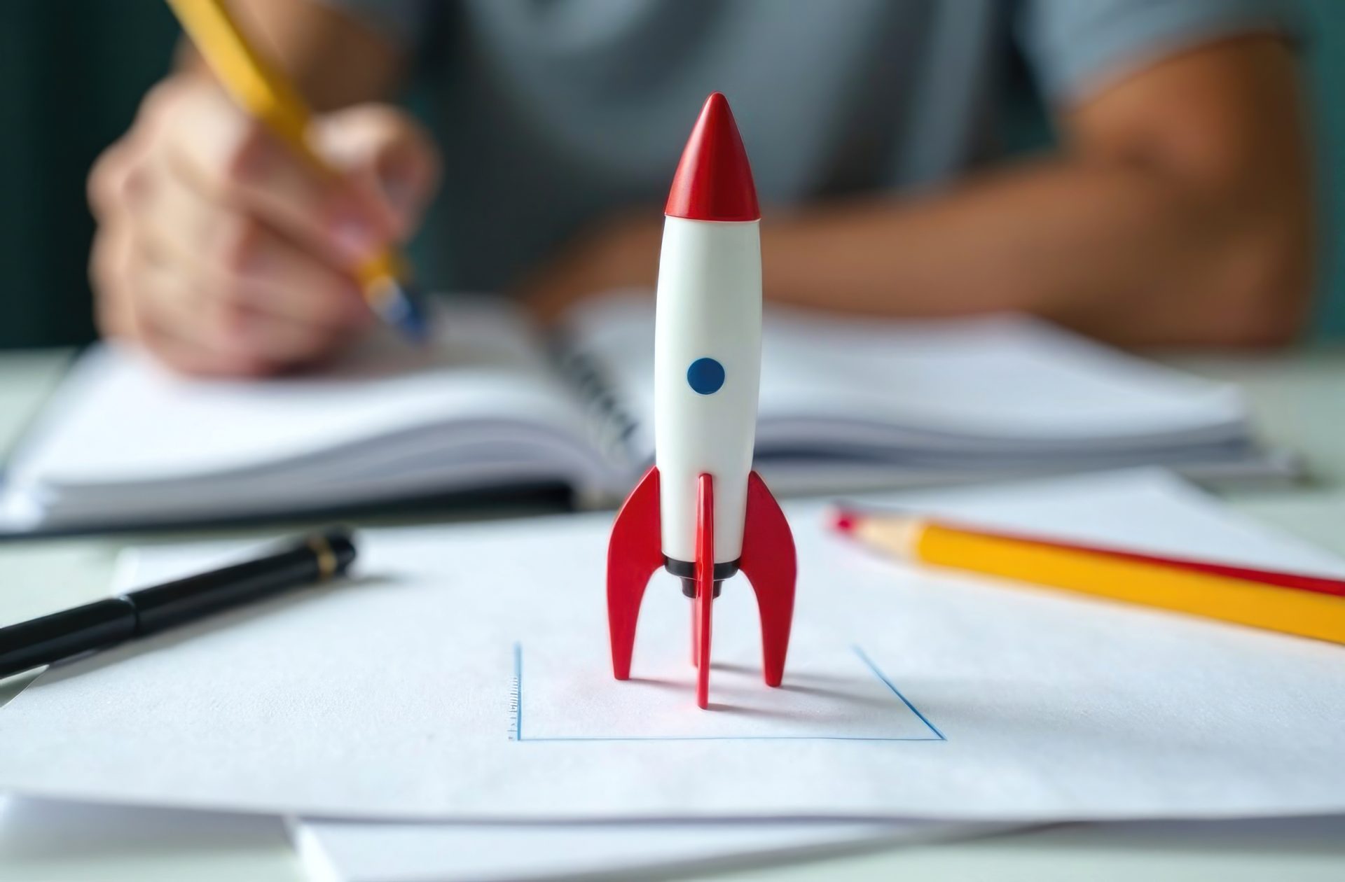 A person is writing in a notebook while a toy rocket captures attention on the table around study materials